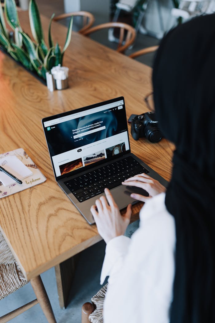 gallery-4 Woman in hijab working on a laptop at a wooden table, camera nearby.