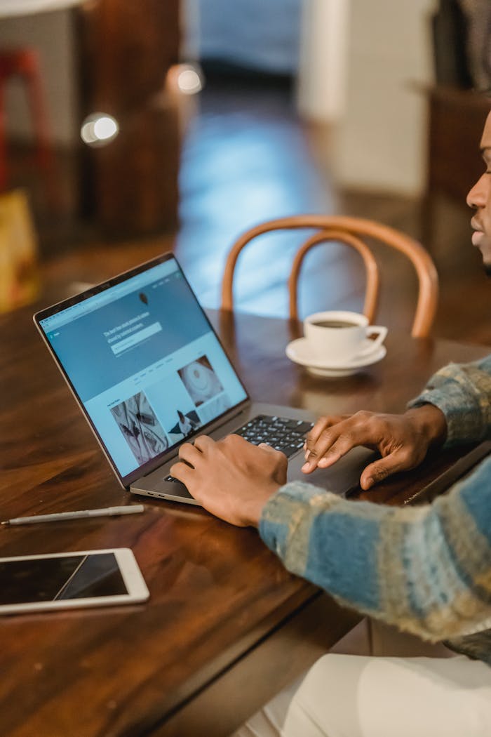 gallery-2 Crop unrecognizable Black male freelancer in casual clothes sitting at wooden table with laptop and typing on keyboard of modern interior